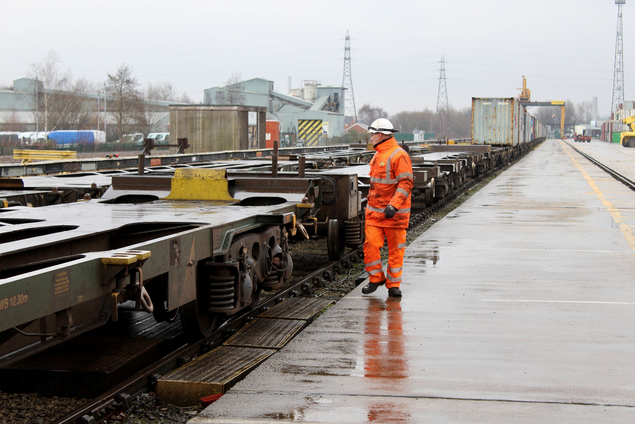 Rail worker in orange PPE inspecting freight wagons on a wet rail terminal.
