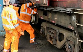 Two rail workers in orange PPE securing a freight container to a wagon.