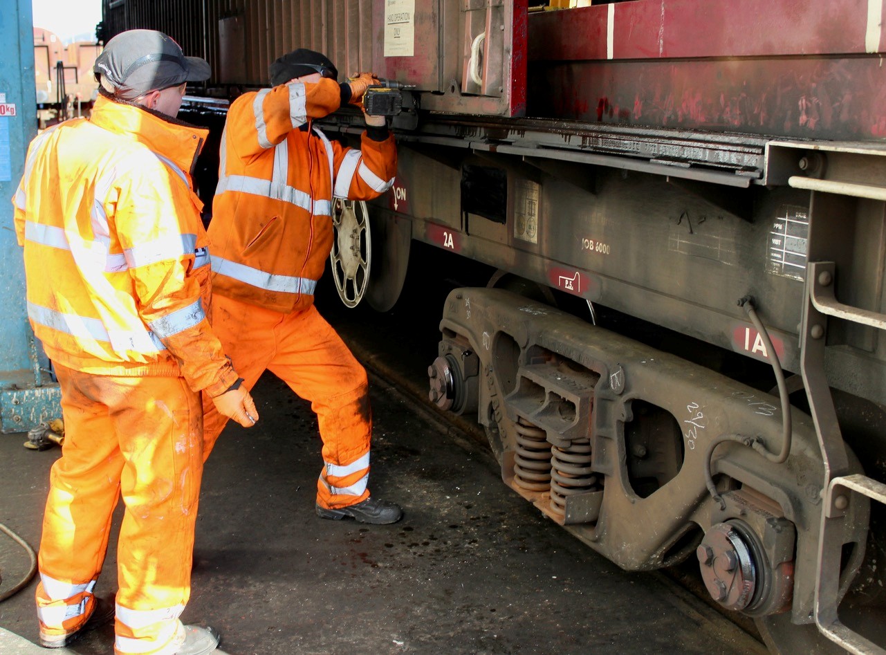 Two rail workers in orange PPE securing a freight container to a wagon.
