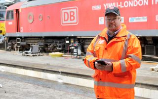 Rail worker in orange high-visability clothing standing in front of a DB locomotive while holding a mobile phone