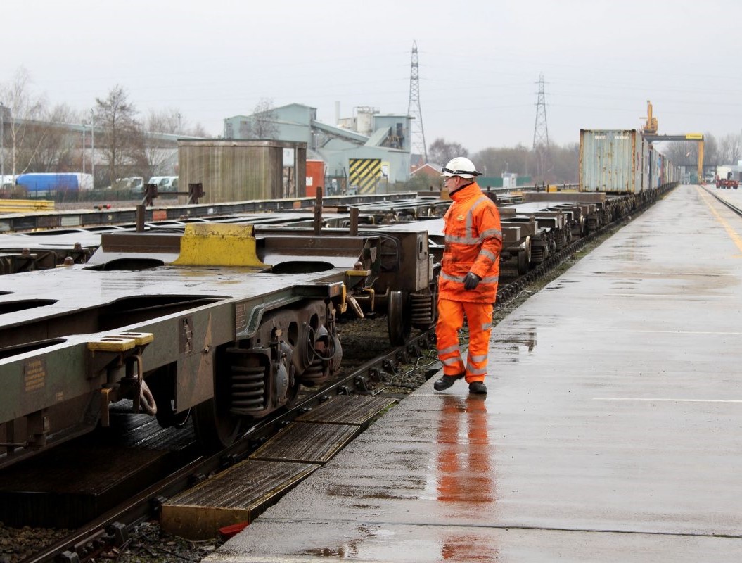 Rail worker in orange PPE inspecting freight wagons on a wet rail terminal